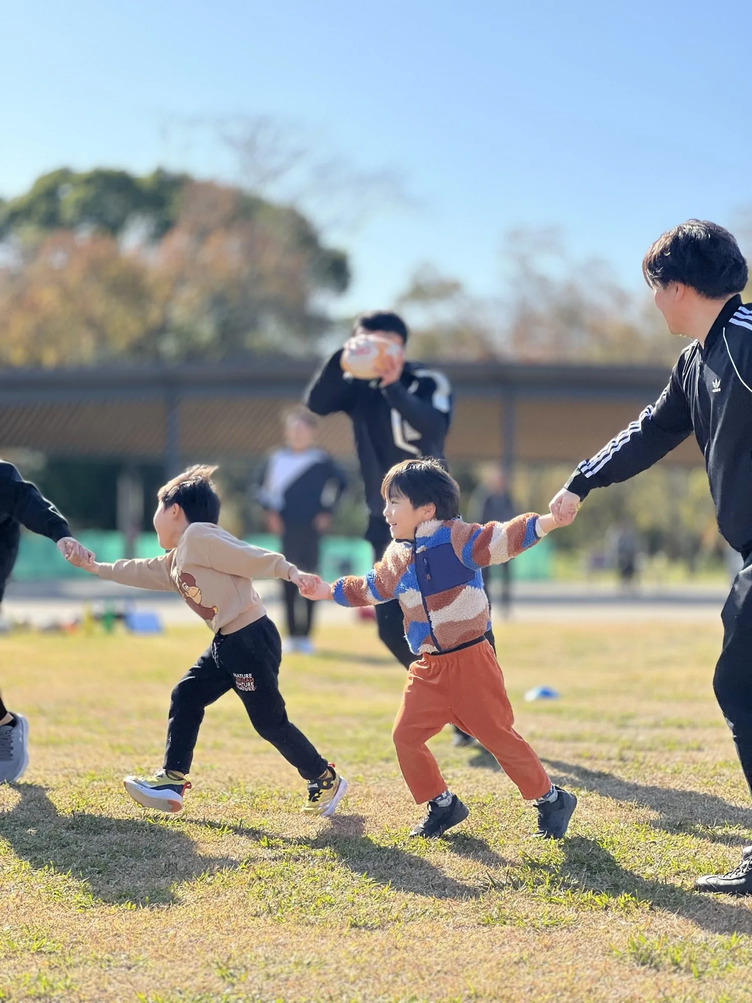 夢の島の芝生で、手をつないで走る子どもたちとコーチ