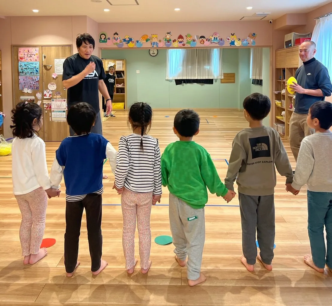 保育園の部屋で、手をつないで一列に並ぶ園児たちとコーチ